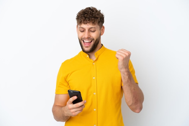 Jonge knappe blanke man geïsoleerd op een witte achtergrond met telefoon in overwinningspositie