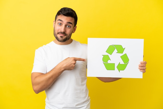 Jonge knappe blanke man geïsoleerd op gele achtergrond met een bordje met recycle icoon en erop wijzend