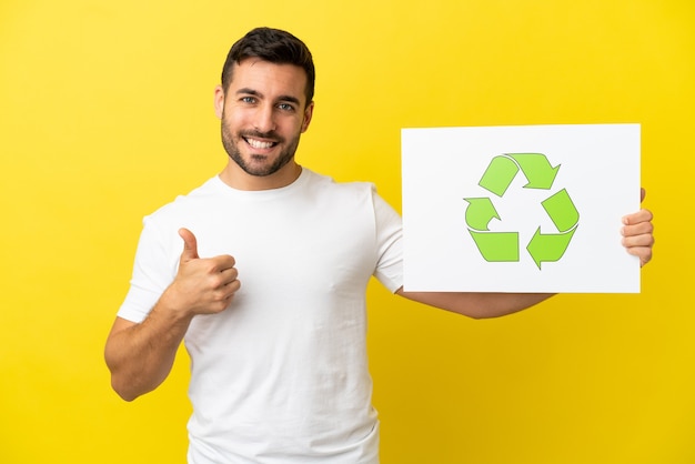 Jonge knappe blanke man geïsoleerd op gele achtergrond met een bordje met recycle pictogram met duim omhoog