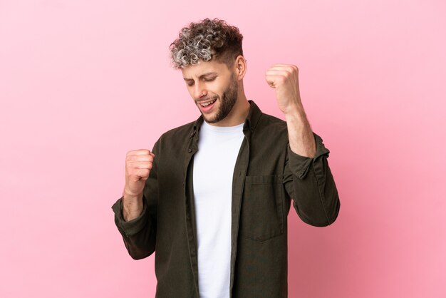 Jonge knappe blanke man geïsoleerd op roze achtergrond die een overwinning viert