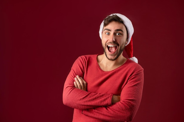 Jonge knappe blanke man in rode trui en kerstman hoeden staat op rode achtergrond in de studio en