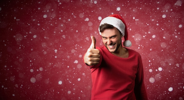Jonge knappe blanke man in rode trui en kerstmuts staat op rode achtergrond in studio duimen opdagen. Close-up portret Europese man met kerstsfeer met sneeuw Vakantie banner.