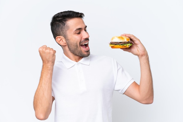 Jonge knappe blanke man met een hamburger over geïsoleerde achtergrond die een overwinning viert