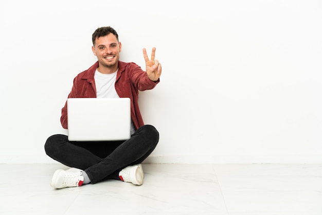 Jonge knappe blanke man sit-in op de vloer met laptop glimlachend en overwinningsteken tonen