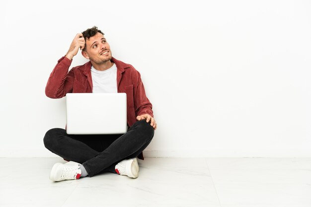 Jonge knappe blanke man sit-in op de vloer met laptop twijfels en met verwarde gezichtsuitdrukking