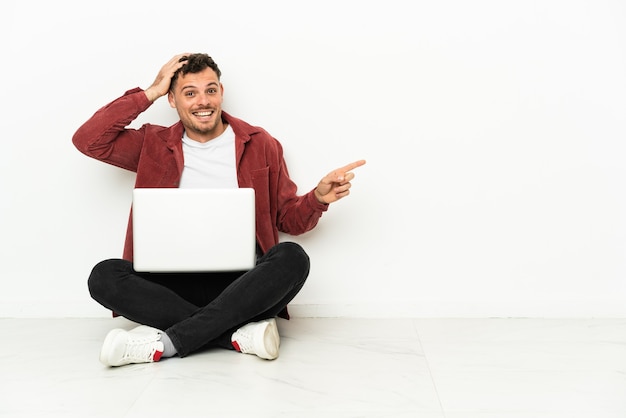 Jonge knappe blanke man sit-in op de vloer met laptop verrast en wijzende vinger naar de zijkant