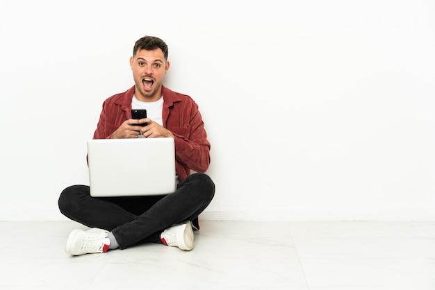 Jonge knappe blanke man zit op de vloer met laptop verrast en stuurt een bericht a