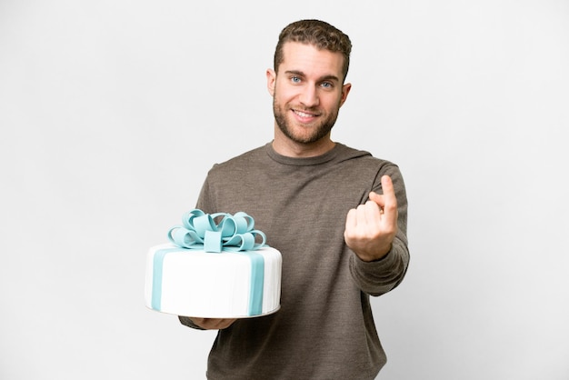 Jonge knappe blonde man met een grote cake over een geïsoleerde witte achtergrond die een komend gebaar doet