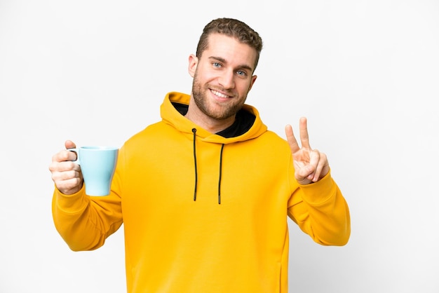 Jonge knappe blonde man met kopje koffie over geïsoleerde achtergrond glimlachend en overwinningsteken tonen
