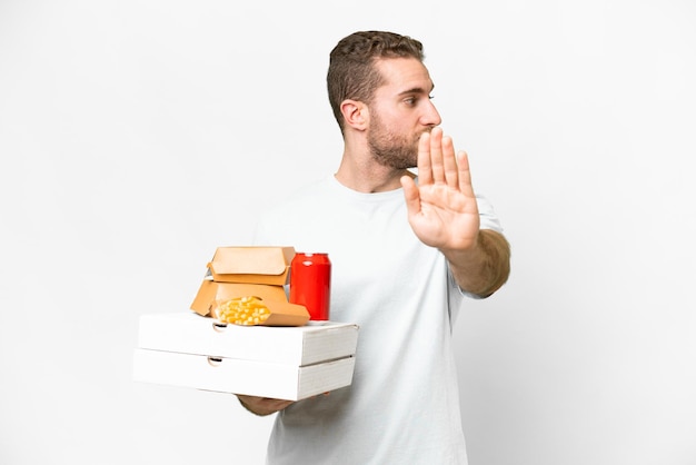 Jonge knappe blonde man met pizza's en hamburgers over geïsoleerde achtergrond stop gebaar maken en teleurgesteld