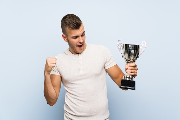 Jonge knappe blondemens over geïsoleerde blauwe achtergrond die een trofee houden