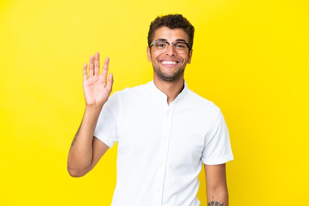 Jonge knappe Braziliaanse man geïsoleerd op gele achtergrond saluerend met de hand met gelukkige uitdrukking