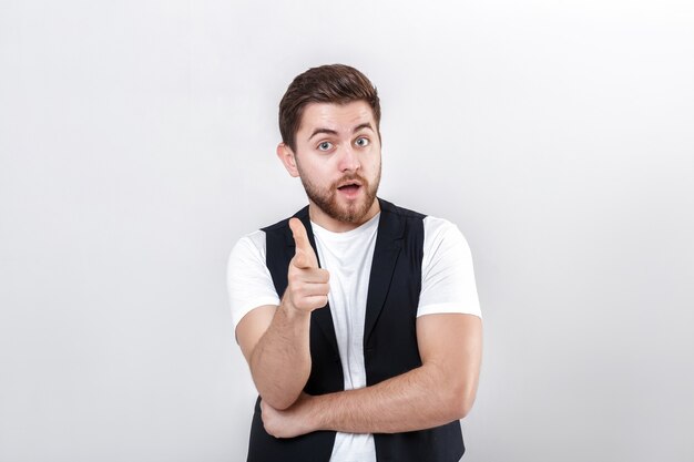 Jonge knappe brunette man met een baard in een wit overhemd en een zwart vest die naar voren wijst en glimlacht