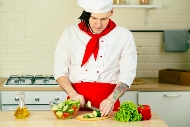 Jonge knappe chef-kok die groene komkommer snijdt op een houten bord in de keuken Positieve man met dreadlocks in uniform die groentesalade bereidt