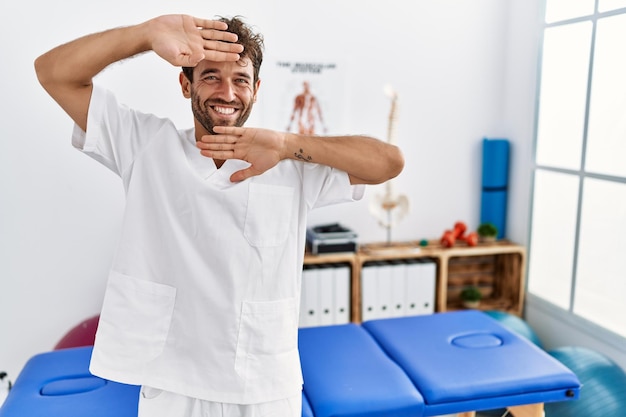 Jonge knappe fysiotherapeut man aan het werk bij pijn herstel kliniek glimlachend vrolijk spelen peek a boo met handen tonen gezicht verrast en opgewonden