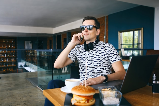 Jonge knappe hipster man in zonnebril zitten in café praten mobiele telefoon. Laptop, kopje koffie en cheeseburger op houten tafel.