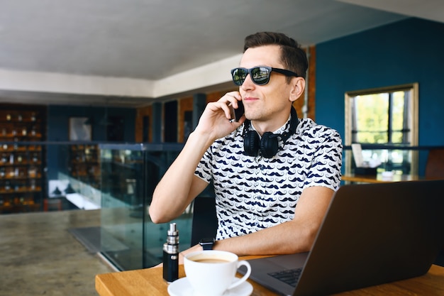 Jonge knappe hipster man in zonnebril zitten in café praten mobiele telefoon. Laptop op houten tafel.