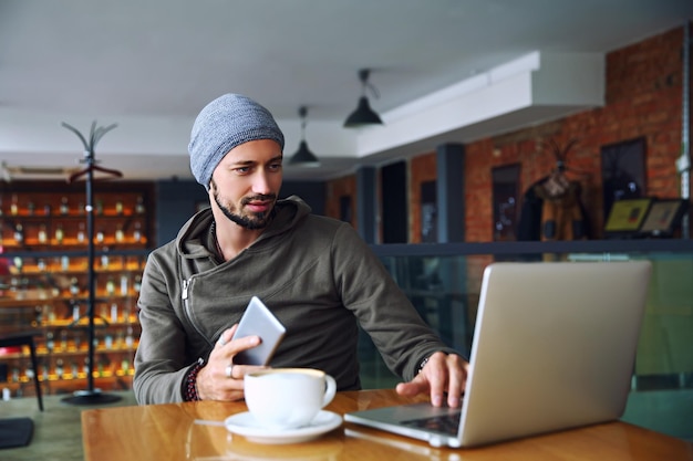 Jonge knappe hipster man met baard gebruikt computer in cafetaria