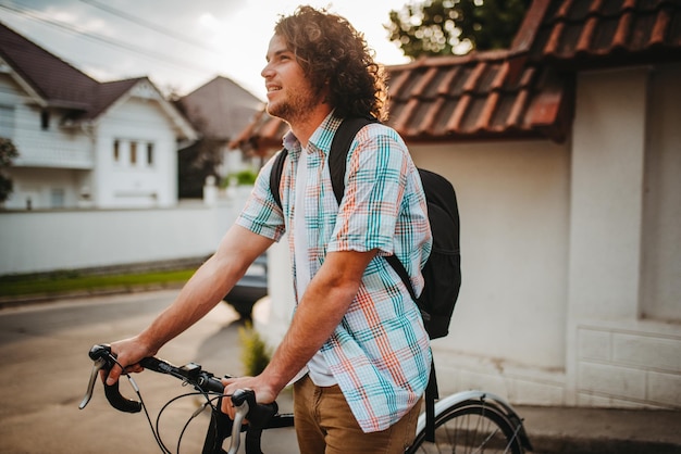 Jonge knappe hipster man met krullend haar met rugzak fietsen op de stadsstraat voor ecologisch leven Man student fietsen met fiets naar het college Mensen levensstijl en sport