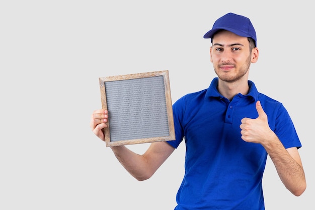 Jonge knappe jongen in bezorguniform die leeg frame vasthoudt en duimen omhoog geeft