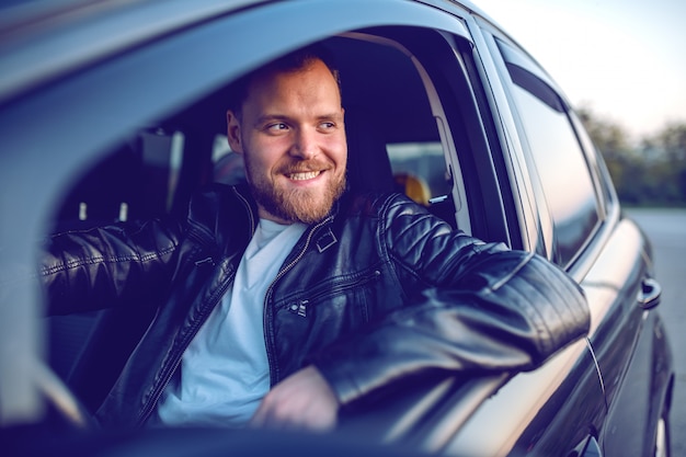 Foto jonge knappe lachende bebaarde blonde blanke man in lederen jas poseren in zijn moderne auto.