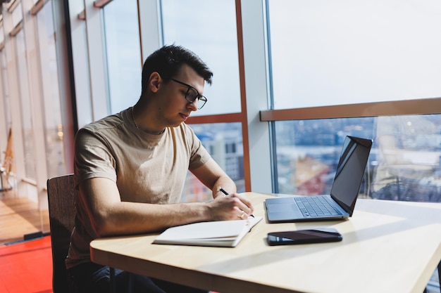 Jonge knappe lachende slimme freelancer in glazen werkt op een laptop terwijl hij tijd doorbrengt in een café Een positieve student zit aan een houten tafel in een café Gelukkige man zit terwijl hij binnenshuis werkt