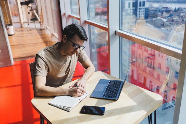 Jonge knappe lachende slimme freelancer in glazen werkt op een laptop terwijl hij tijd doorbrengt in een café Een positieve student zit aan een houten tafel in een café Gelukkige man zit terwijl hij binnenshuis werkt