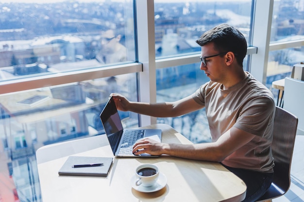 Jonge knappe lachende slimme freelancer in glazen werkt op een laptop terwijl hij tijd doorbrengt in een café Een positieve student zit aan een houten tafel in een café Gelukkige man zit terwijl hij binnenshuis werkt