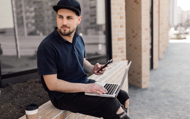 Jonge knappe man aan het werk met laptop en smartphone online op straat