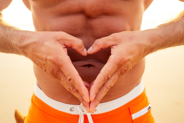 Jonge knappe man crème zonnebrand lotion toe te passen op het strand van de zee. sexy mannelijk model atleet bodybuilder poseren het perfecte atletische lichaam, epileren en ontharen shugaring concept. hart vorm handen.