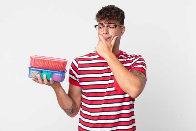 Jonge knappe man denkt, voelt zich twijfelachtig en verward en houdt een lunchdoos vast