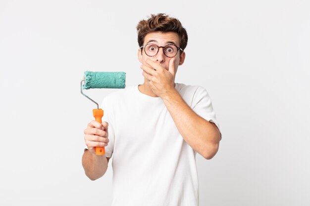 Jonge knappe man die de mond bedekt met handen met een geschokte en een verfroller vasthoudt