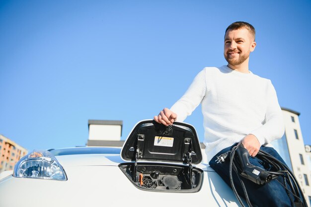 Jonge knappe man die een oplaadkabel vasthoudt op het punt van een elektrisch laadstation in de buurt van zijn nieuwe auto.