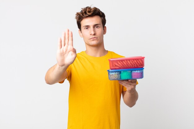 jonge knappe man die er serieus uitziet met een open palm die een stopgebaar maakt en lunchboxen vasthoudt