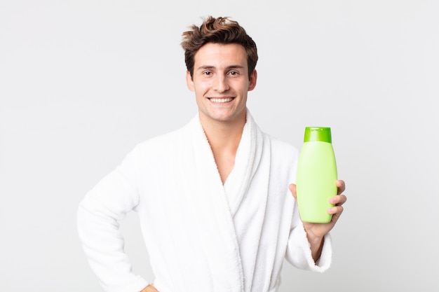 Jonge knappe man die gelukkig lacht met een hand op de heup en zelfverzekerd met een badjas en een shampoofles vasthoudt