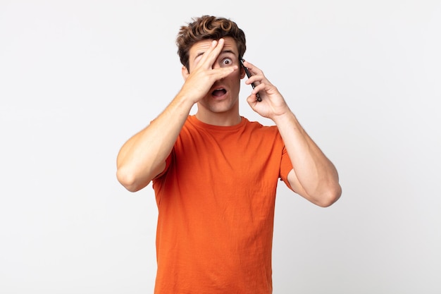 Foto jonge knappe man die geschokt, bang of doodsbang kijkt, zijn gezicht bedekt met de hand en praat met een smartphone