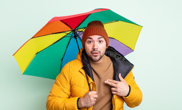 Jonge knappe man die geschokt en verrast kijkt met wijd open mond, wijzend naar zichzelf. regen en paraplu concept