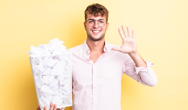 Jonge knappe man die lacht en er vriendelijk uitziet, met nummer vijf. papier ballen prullenbak concept