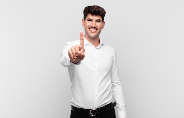 Jonge knappe man die lacht en er vriendelijk uitziet, nummer één toont of eerst met de hand naar voren, aftellend