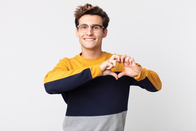 Jonge knappe man die lacht en zich gelukkig, schattig, romantisch en verliefd voelt, en met beide handen hartvorm maakt