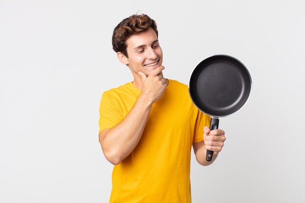 Jonge knappe man die lacht met een gelukkige, zelfverzekerde uitdrukking met de hand op de kin en een kookpan vasthoudt