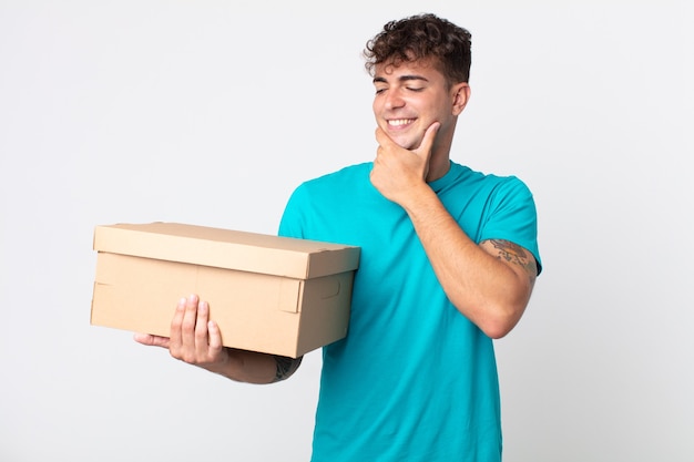 Jonge knappe man die lacht met een vrolijke, zelfverzekerde uitdrukking met de hand op de kin en een kartonnen doos vasthoudt