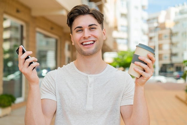 Jonge knappe man die mobiele telefoon gebruikt en een kopje koffie vasthoudt met een gelukkige uitdrukking