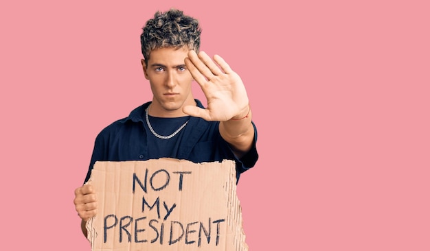 Jonge knappe man die niet mijn president-protestbanner vasthoudt met open hand die stopbord doet met serieuze en zelfverzekerde uitdrukking, verdedigingsgebaar