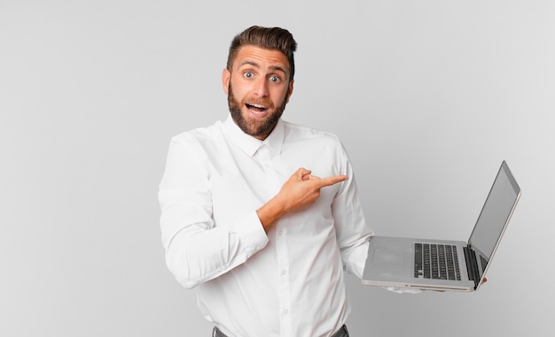 Jonge knappe man die opgewonden en verrast kijkt en naar de zijkant wijst en een laptop vasthoudt