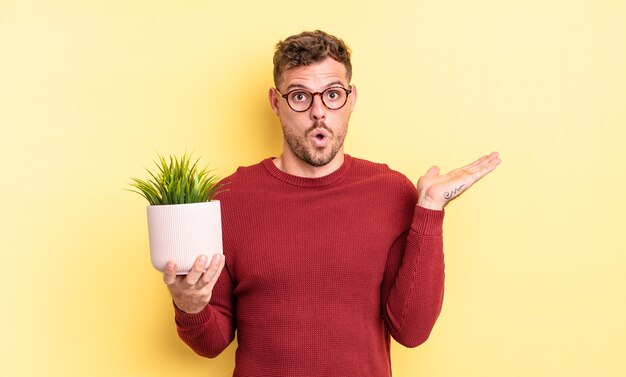 Jonge knappe man die verrast en geschokt kijkt, met open mond terwijl hij een object vasthoudt. decoratief plantenconcept