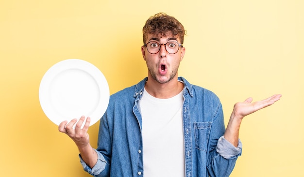 Jonge knappe man die verrast en geschokt kijkt, met open mond terwijl hij een voorwerp vasthoudt. leeg schotelconcept