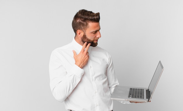 Jonge knappe man die vrolijk lacht en dagdroomt of twijfelt en een laptop vasthoudt