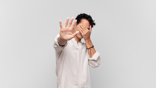 Jonge knappe man die zijn gezicht bedekt met de hand en de andere hand naar voren steekt om de camera te stoppen, foto's of afbeeldingen te weigeren