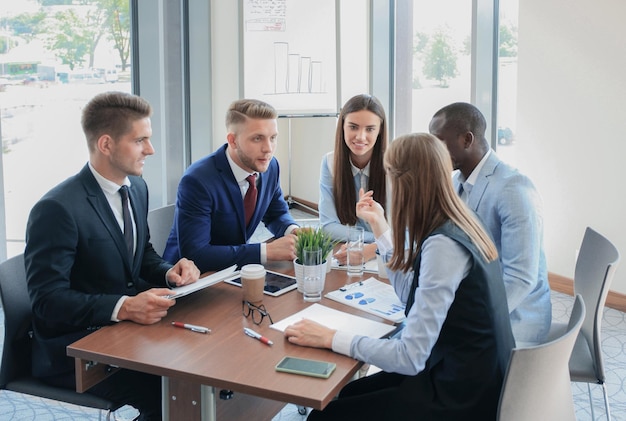 Jonge knappe man gebaren en iets bespreken terwijl zijn collega's naar hem luisteren terwijl hij aan de kantoortafel zit